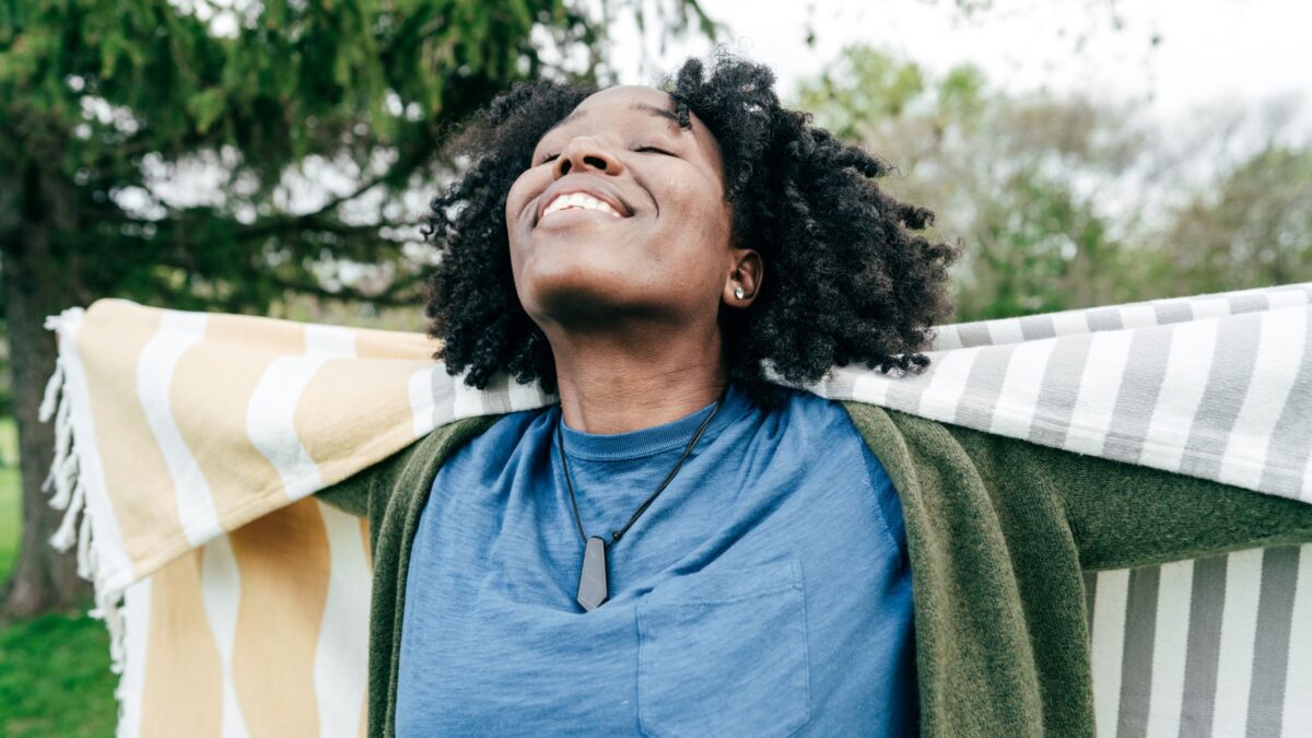 A black woman practicing self-care in the park, holding a towel and embracing her wellbeing with arms outstretched.